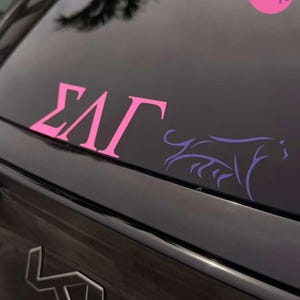 May include: A pink vinyl decal featuring the Greek letters Sigma, Lambda, and Gamma applied to a dark car window. To the right of the letters is a purple line-art illustration of a panther. A circular pink logo with a panther and the text The Gamma Label is visible in the top right corner.
