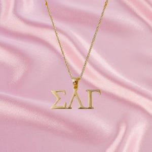 Puede incluir: Un collar de tono dorado con un colgante que presenta las letras griegas Sigma, Lambda y Gamma. El colgante está suspendido de una delicada cadena de tono dorado. Las letras están recortadas con un acabado metálico limpio, creando un símbolo distintivo y reconocible para la organización de la hermandad.