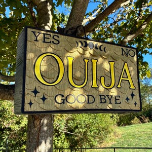 May include: A rectangular wooden Ouija board sign with the words "YES" and "NO" at the top, and "OUIJA GOOD BYE" in yellow lettering. The sign is attached to a tree branch, with a natural wood grain background and black trim.
