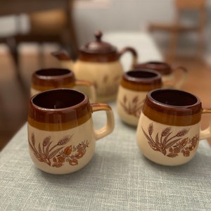 May include: A set of brown and beige ceramic mugs with a wheat and floral design. The mugs are arranged on a light blue tablecloth with a teapot in the background.