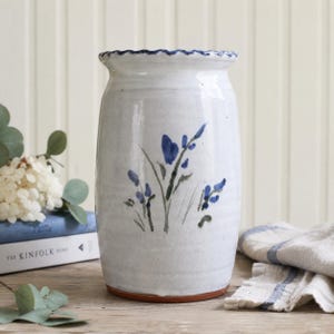 May include: A cylindrical ceramic vase with a glossy off-white glaze and a scalloped rim. The exterior features a hand-painted botanical design in shades of blue and green, depicting delicate stems and small flower buds centered on the body.