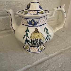 May include: White ceramic teapot with a blue, green, and yellow floral design. The teapot has a lid with a knob on top. The teapot is sitting on a white brick surface.
