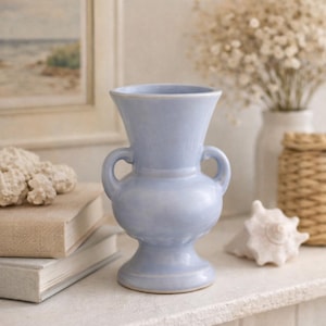 May include: A light blue ceramic vase with two handles. The vase has a bulbous body and a flared rim. It is displayed on a white surface with other decorative items, including books and a seashell. The vase is a decorative home accent.