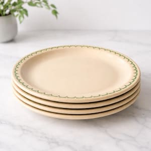 May include: A stack of four cream-colored ceramic plates. Each plate has a scalloped green border. The plates are stacked on top of each other, with the top plate in focus. The background is a marble surface.