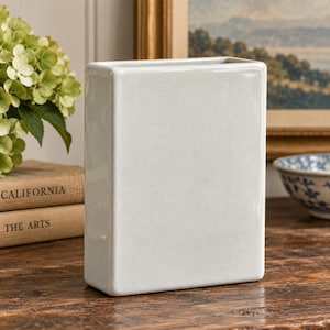 May include: A rectangular ceramic vase with a smooth, glossy, off-white finish. The vase features a flat front and back with rounded vertical edges, creating a clean, boxy silhouette. The top opening is a narrow, elongated rectangle shape.