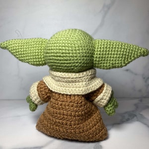 May include: A crocheted green and brown plush toy with large ears. The toy is wearing a brown robe with a white collar.