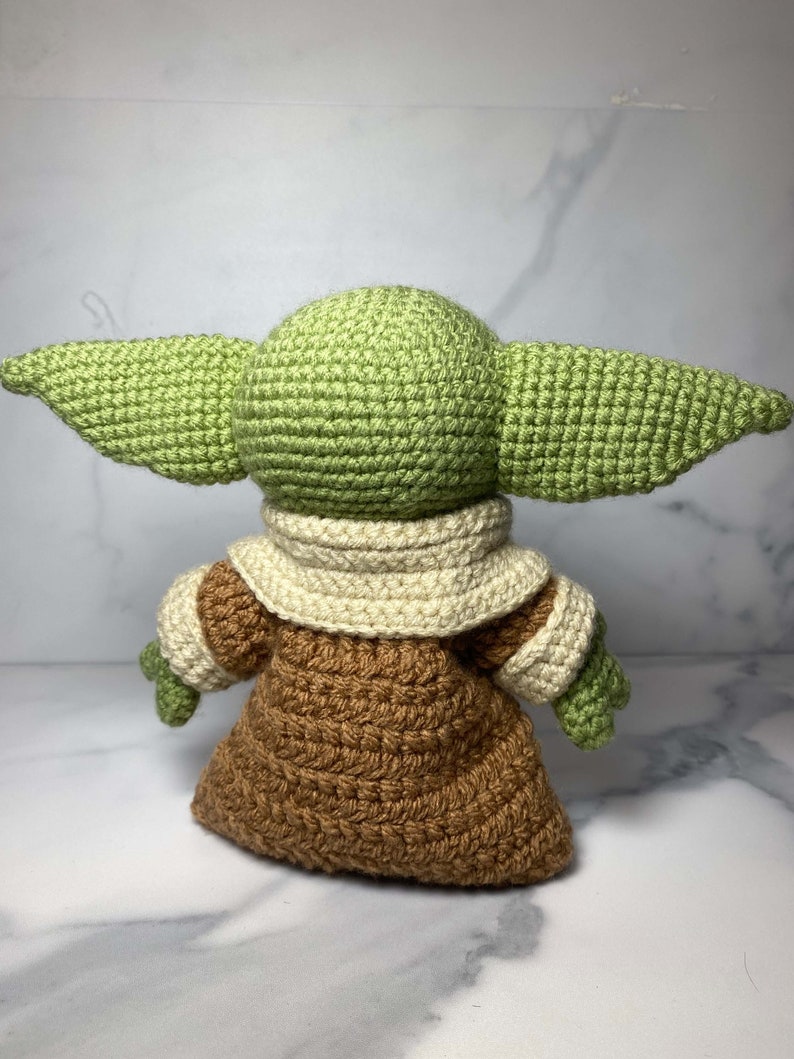 May include: A crocheted green and brown plush toy with large ears. The toy is wearing a brown robe with a white collar.