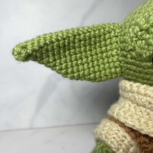 May include: A green crocheted plush toy with a large, pointed ear. The toy is wearing a cream-colored crocheted collar and has brown crocheted details on its arms.