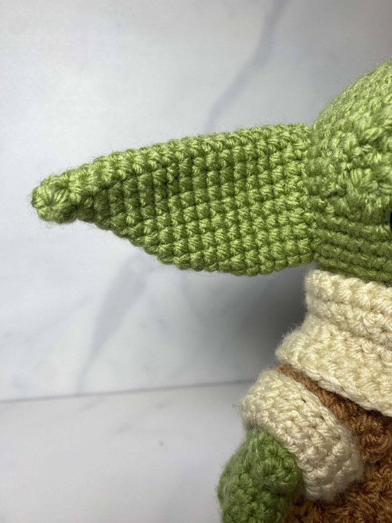 May include: A green crocheted plush toy with a large, pointed ear. The toy is wearing a cream-colored crocheted collar and has brown crocheted details on its arms.