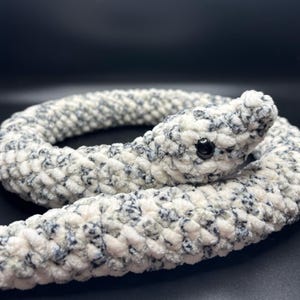 Op de afbeelding: Een wit en grijs gehaakt slangen knuffeldier met zwarte ogen. De slang is opgerold in een cirkel.