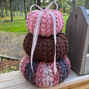 May include: A stacked, handmade pumpkin decoration. The top pumpkin is pink, the middle is brown, and the bottom is a mix of pink, gray, and red. A white lace ribbon is tied around the top pumpkin. The pumpkins are made of a textured yarn.