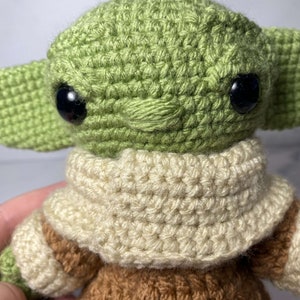 May include: A crocheted green and brown plush toy with large black eyes and a white crocheted scarf. The toy is a popular character from the Star Wars franchise.