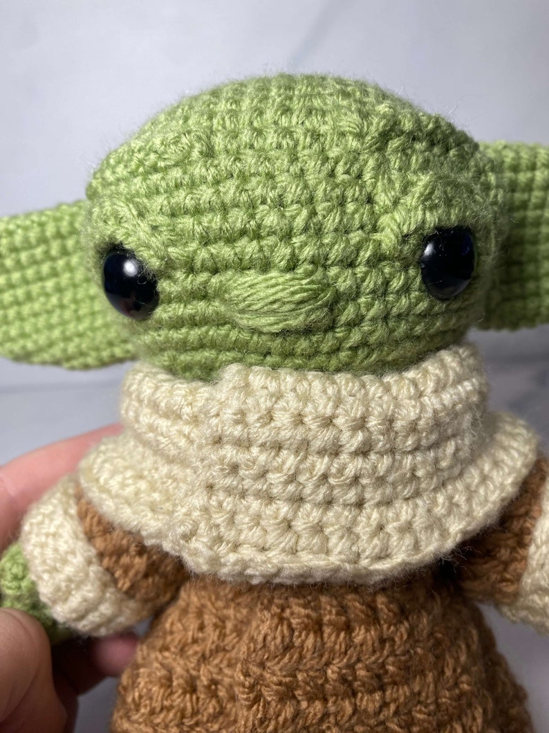 May include: A crocheted green and brown plush toy with large black eyes and a white crocheted scarf. The toy is a popular character from the Star Wars franchise.