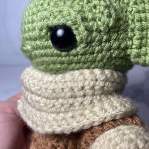 May include: A crocheted green and brown plush toy with a white scarf. The toy has large black eyes and a small mouth.