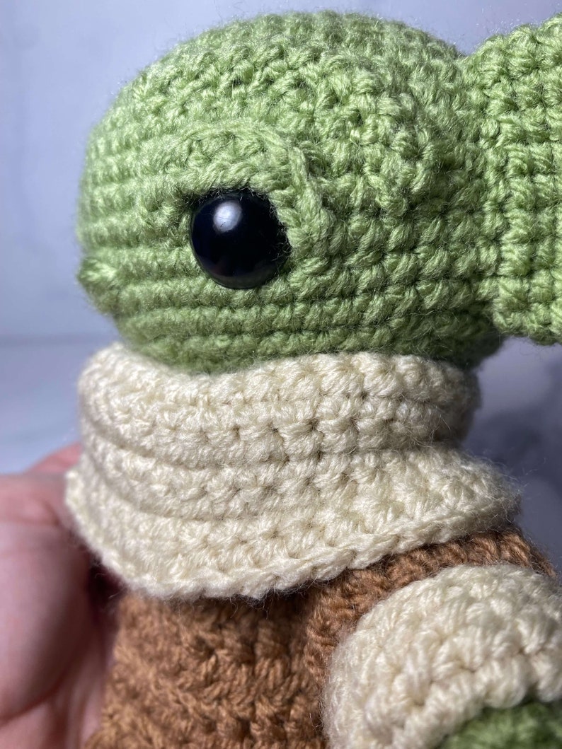 May include: A crocheted green and brown plush toy with a white scarf. The toy has large black eyes and a small mouth.