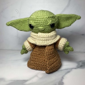 May include: A crocheted green and brown Yoda doll with large black eyes. The doll is wearing a brown robe with a white collar.