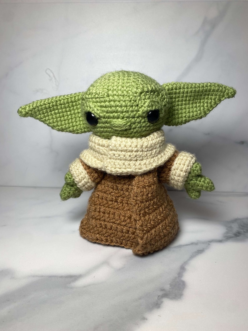 May include: A crocheted green and brown Yoda doll with large black eyes. The doll is wearing a brown robe with a white collar.