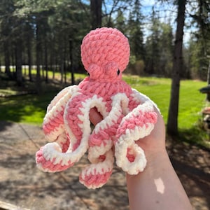 Może przedstawiać: Ręcznie robiona ośmiornica na szydełku w odcieniach różu i bieli. Ośmiornica ma okrągłą głowę i osiem zakręconych macek. Macki są w paski w kolorze różowym i białym. Ośmiornica to miękka, pluszowa zabawka.