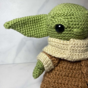 May include: A green crocheted plush toy with a brown crocheted coat and a white crocheted scarf. The toy has large black eyes and a pointed ear.
