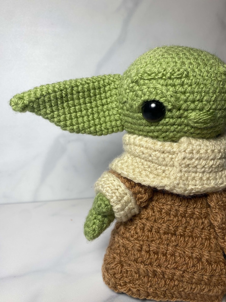 May include: A green crocheted plush toy with a brown crocheted coat and a white crocheted scarf. The toy has large black eyes and a pointed ear.
