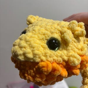 May include: Close-up of a crocheted stuffed animal with yellow and orange fur. The animal has large, black eyes with green pupils.