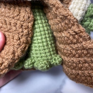 May include: A crocheted green and brown doll wearing a brown coat with a white crocheted collar.