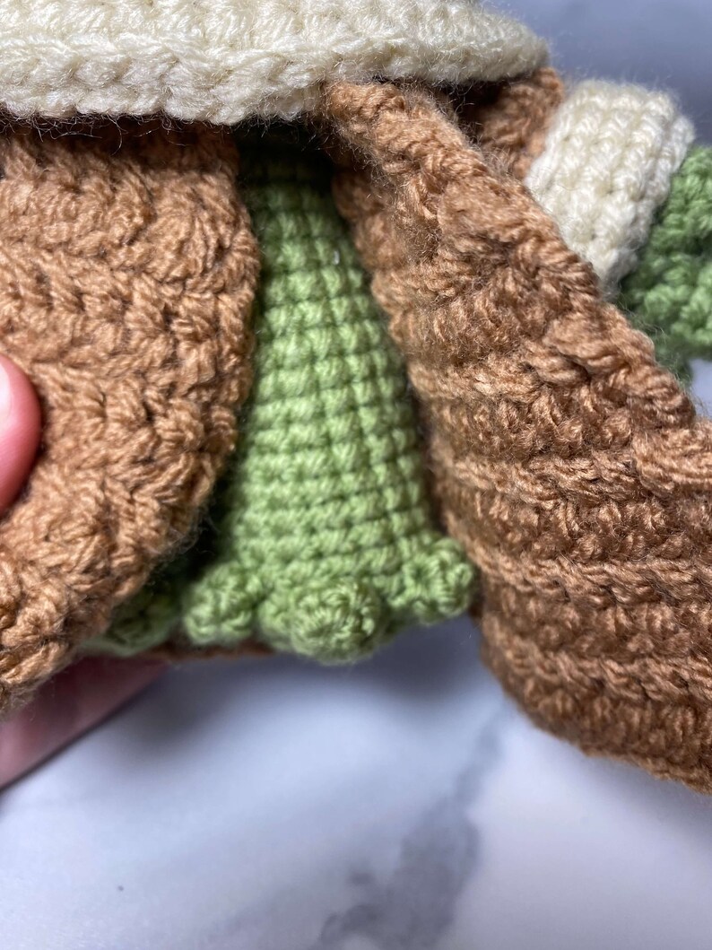 May include: A crocheted green and brown doll wearing a brown coat with a white crocheted collar.
