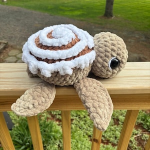 Puede incluir: Una tortuga de peluche hecha a mano con un caparazón de rollo de canela. El caparazón de la tortuga es marrón y blanco, parecido a un rollo de canela. La tortuga tiene aletas, cabeza y un ojo negro. El peluche está sobre una superficie de madera.