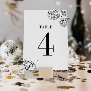 Puede incluir: Tarjeta blanca con texto negro que dice "TABLE 4" y un diseño de bola de discoteca en blanco y negro. La tarjeta está de pie sobre un bloque de madera en una mesa cubierta de confeti plateado y dorado.
