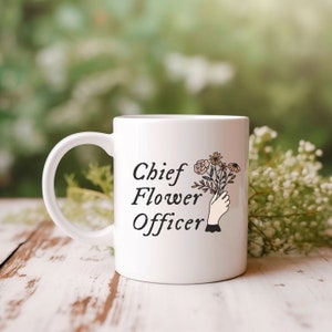 May include: White ceramic mug with a black design that says "Chief Flower Officer" and a hand holding a bouquet of flowers.