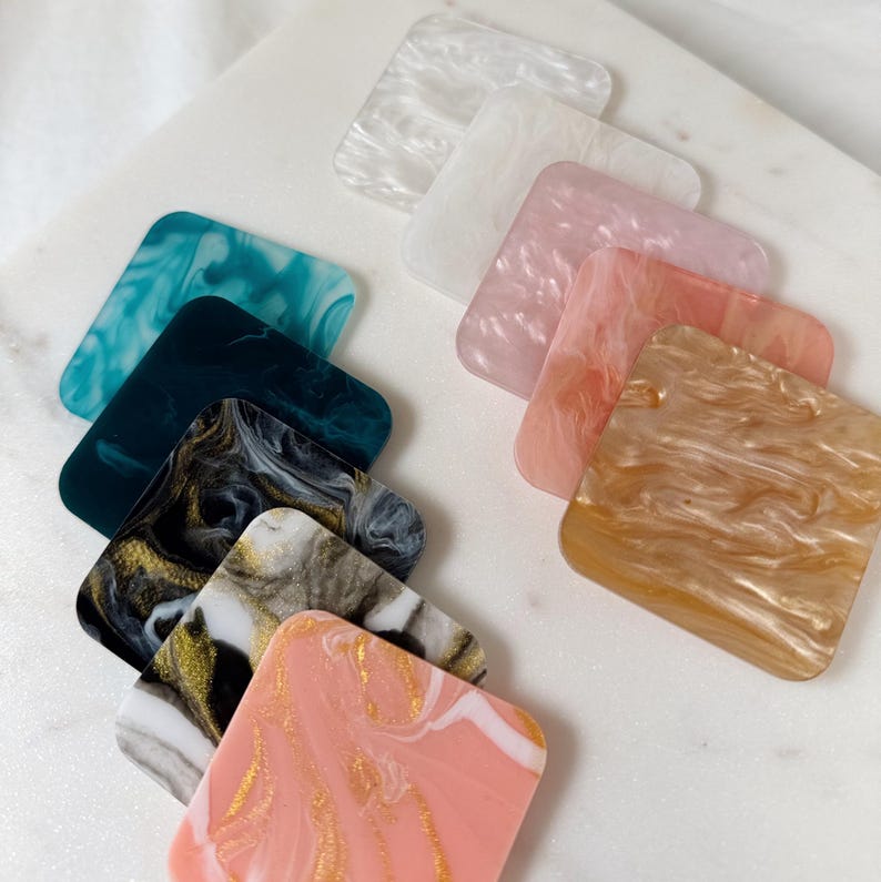 May include: A set of ten square coasters with different marbled patterns. The coasters are made of a translucent material with swirls of white, pink, gold, black, and teal.