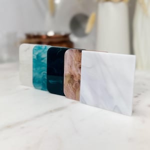May include: A set of five square coasters arranged in a row, featuring distinct marbled patterns in white, turquoise, dark green, peach, and a light grey with white veining, all crafted from a smooth, polished acrylic material with rounded corners.