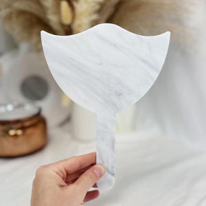 Puede incluir: Espejo de mano con dise&ntilde;o de m&aacute;rmol blanco y gris, con una forma &uacute;nica de escudo. El espejo tiene un mango largo y est&aacute; sostenido por una mano. El fondo es borroso, con elementos decorativos.