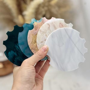 May include: A set of five circular coasters with scalloped edges, featuring a marbled resin finish in shades of dark green, teal, peach with gold flecks, cream, and white with grey veining, arranged in a fanned-out stack for display.