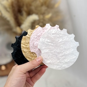 May include: A collection of four decorative coasters in black, gold, pink, and white. Each coaster has a scalloped edge and a swirled, marbled pattern. The coasters are held in a hand, showcasing their varying colors and textures.