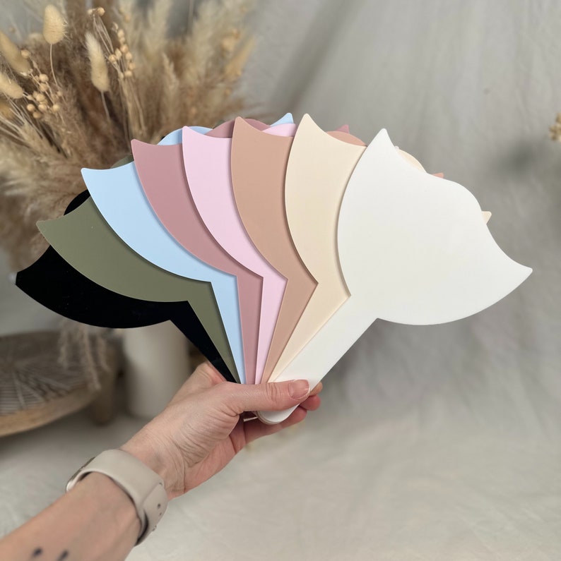 Puede incluir: Una colecci&oacute;n de espejos de mano en varios colores pastel, incluyendo negro, verde oliva, azul claro, malva, rosa, beige y crema. Cada espejo tiene una forma estilizada &uacute;nica con un mango, sostenido en una mano.
