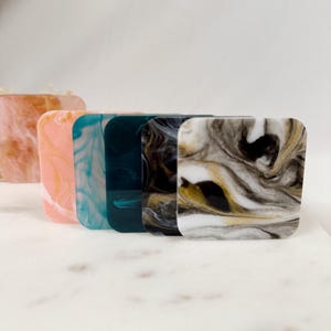 May include: Five square-shaped coasters with a marbled design. The coasters are in various colors, including pink, blue, green, black, and white.
