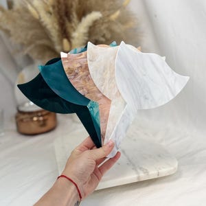 Puede incluir: Una mano sostiene un abanico de tablas de servir decorativas de acr&iacute;lico en varios colores y patrones. Las tablas incluyen tonos de color turquesa, m&aacute;rmol y un dise&ntilde;o de vetas de madera. Las tablas tienen forma de abanico con un asa.