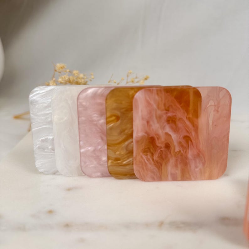 May include: Five square-shaped, translucent, decorative tiles in various shades of pink, white, and gold. The tiles have a marbled or swirled pattern.