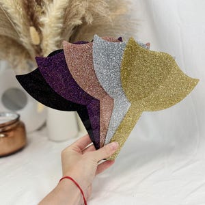 Puede incluir: Espejos de mano con purpurina en varios colores: negro, morado, oro rosa, plateado y dorado. Los espejos tienen una forma estilizada y se sostienen en la mano sobre un fondo suave y neutro.