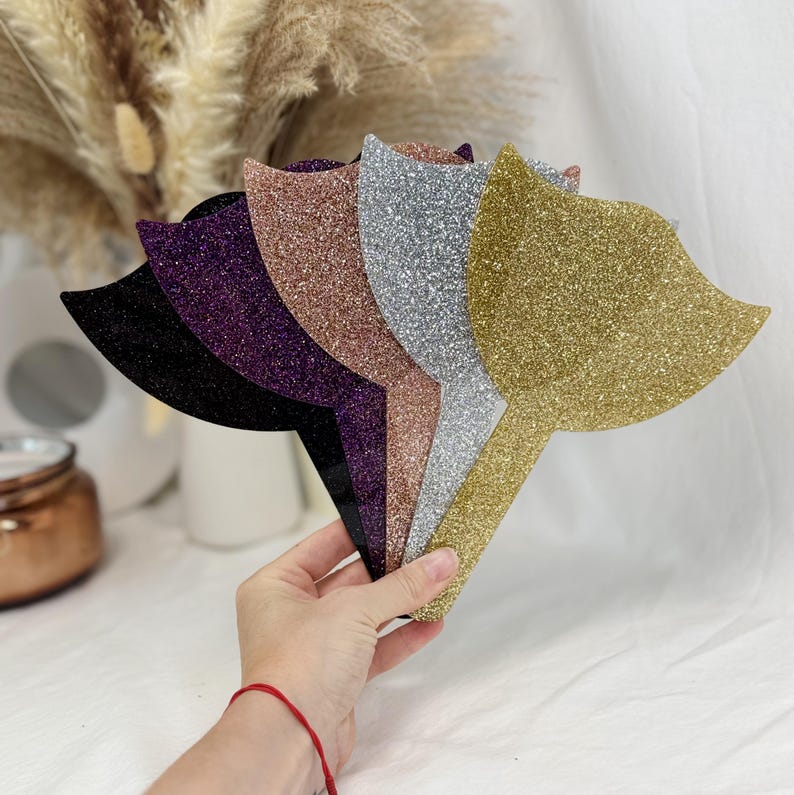 Puede incluir: Espejos de mano con purpurina en varios colores: negro, morado, oro rosa, plateado y dorado. Los espejos tienen una forma estilizada y se sostienen en la mano sobre un fondo suave y neutro.