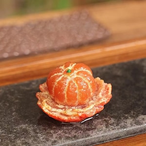 May include: A detailed close-up of a small, orange, mandarin orange fruit, with a green accent, sitting on its peeled rind. The fruit is on a dark, textured surface, with a blurred wooden background.