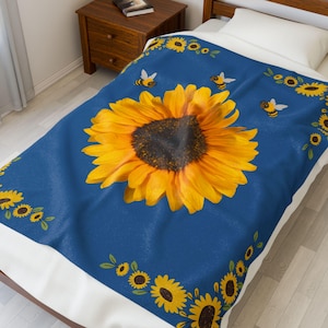 Könnte beinhalten: Eine blaue Decke mit einer großen gelben Sonnenblume und kleineren Sonnenblumen am Rand. Es gibt auch kleine gelbe Bienen, die um die Sonnenblumen herumfliegen.