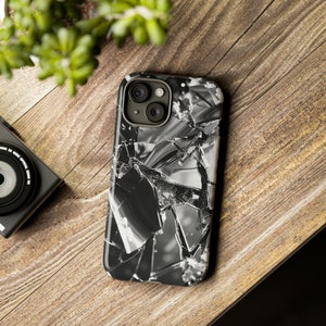 May include: A black and white phone case featuring a shattered glass effect. The case is placed on a wooden surface, with a camera and foliage visible. The case has a glossy finish and a contemporary design.