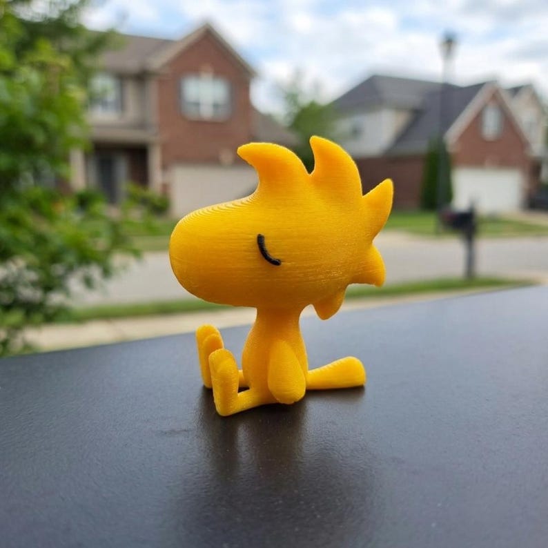 May include: A small, bright yellow 3D-printed figurine of a cartoon bird with a spiky crest and a single black line for a closed eye. The character is depicted in a seated position with its legs extended forward and a small tail visible behind.
