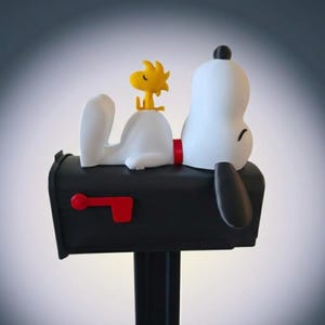 May include: A black mailbox featuring a white dog figure lying on top with a small yellow bird perched on its back. The mailbox has a red flag on the side and a black post. The dog has a black ear and a red collar, while the bird is bright yellow.