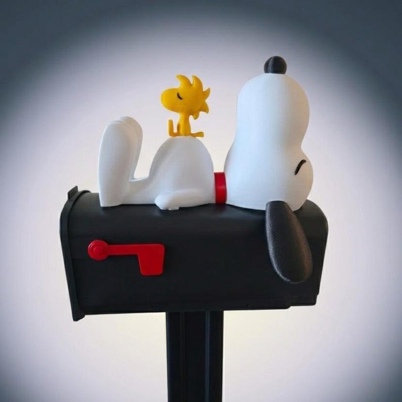 May include: A black mailbox featuring a white dog figure lying on top with a small yellow bird perched on its back. The mailbox has a red flag on the side and a black post. The dog has a black ear and a red collar, while the bird is bright yellow.