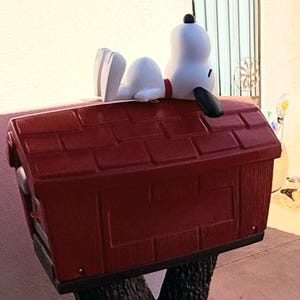 May include: A red mailbox shaped like a doghouse with a textured brick pattern on the sides and a shingled roof. A white Snoopy figure with a black ear and a red collar is lying on its back on top of the roof, facing toward the right side.
