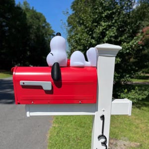 May include: A bright red mailbox mounted on a white post with the number 13 visible. A white Snoopy figure with black ears is positioned on top of the mailbox, lying down in a relaxed pose. The mailbox features a gray flag on the side.