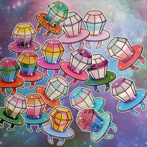 Puede incluir: Un conjunto de 13 pegatinas brillantes, de forma de anillo y con un diseño de diamante. Cada pegatina tiene una combinación de colores diferente y un acabado brillante y holográfico.